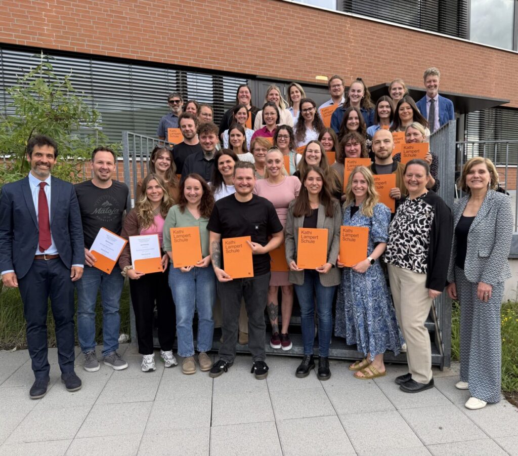 Die Absolvent:innen stehen auf einer Außentreppe vor einem modernen Backsteingebäude und halten orange Mappen mit der Aufschrift „Kathi Lampert Schule“, während die Gruppe für ein gemeinsames Foto posiert. Landesrätin Schöbi-Fink steht auf der rechten Seite.