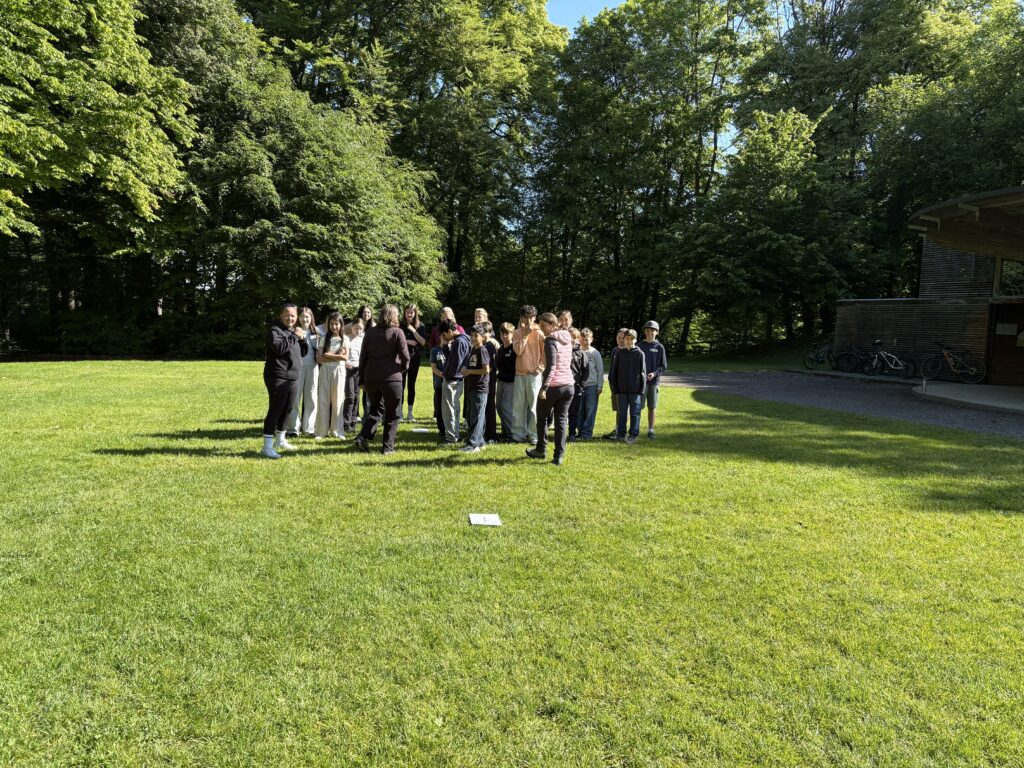 Eine Gruppe von Jugendlichen und Erwachsenen steht gemeinsam auf einer sonnigen Wiese in einem Park und nimmt an einer Aktivität teil. Im Hintergrund sind dichte, grüne Bäume zu sehen.
