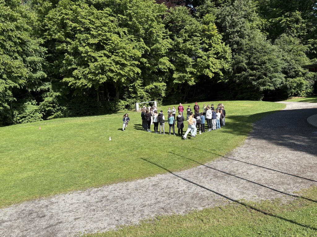 Eine Gruppe von Jugendlichen und Erwachsenen steht im Kreis auf einer Wiese im Park und nimmt an einem gemeinsamen Spiel teil.