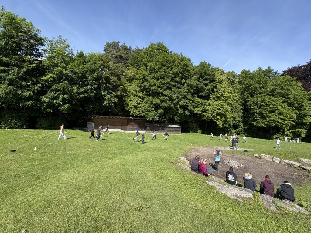 Kinder laufen und spielen auf einer großen Wiese unter blauem Himmel vor einem Wald, im Hintergrund steht ein Holzgebäude.