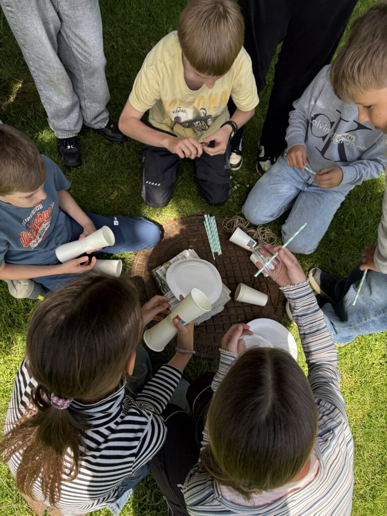 Kinder sitzen im Kreis auf einer Wiese und basteln mit Bechern und Strohhalmen.
