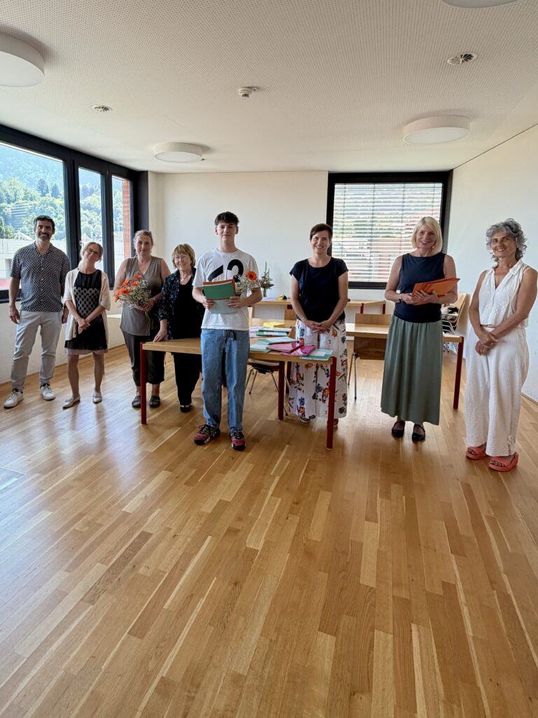 Ein Studierender steht mit dem Zeugnis und Blumen in der Hand vor einem Tisch. Neben ihm stehen insgesamt 7 Personen (Lehrpersonen und Prüfungskommission).