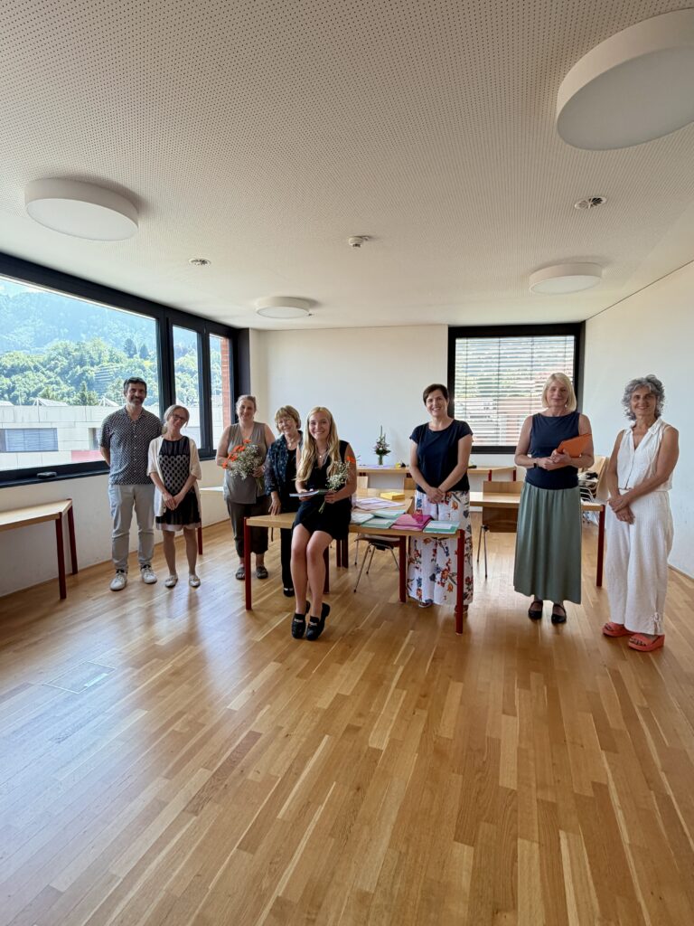 Eine Studierende steht mit dem Zeugnis und Blumen in der Hand vor einem Tisch. Neben ihr stehen insgesamt 7 Personen (Lehrpersonen und Prüfungskommission).