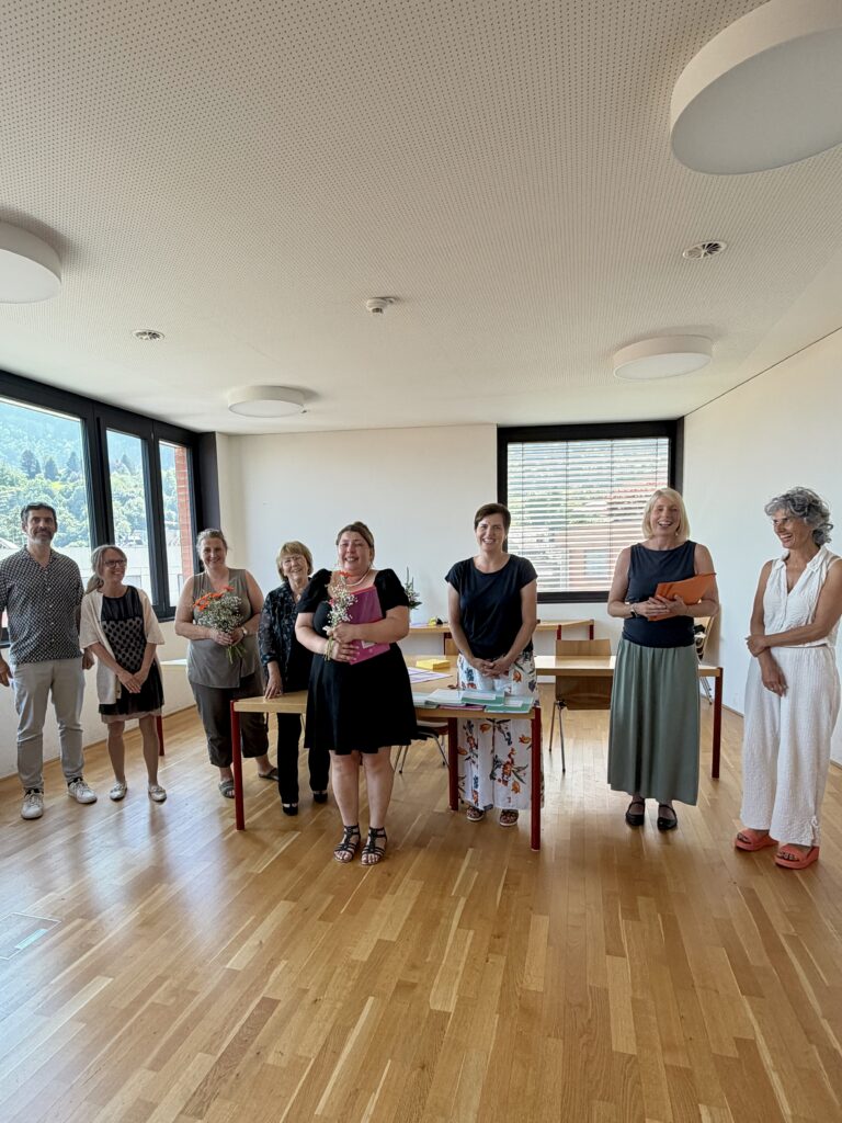Eine Studierende steht mit dem Zeugnis und Blumen in der Hand vor einem Tisch. Neben ihr stehen insgesamt 7 Personen (Lehrpersonen und Prüfungskommission).