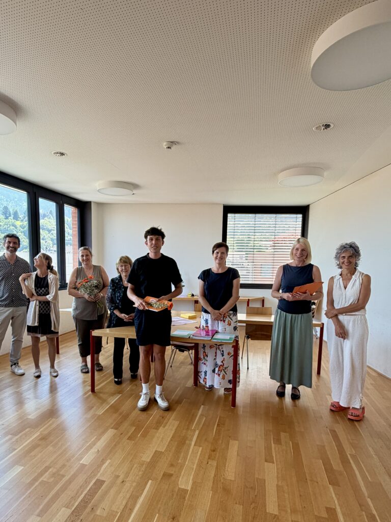 Ein Studierender steht mit dem Zeugnis und Blumen in der Hand vor einem Tisch. Neben ihm stehen insgesamt 7 Personen (Lehrpersonen und Prüfungskommission)