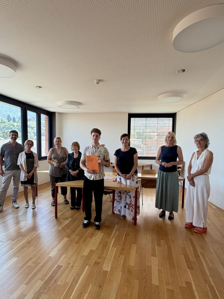 Ein Studierender steht mit dem Zeugnis und Blumen in der Hand vor einem Tisch. Neben ihm stehen insgesamt 7 Personen (Lehrpersonen und Prüfungskommission).