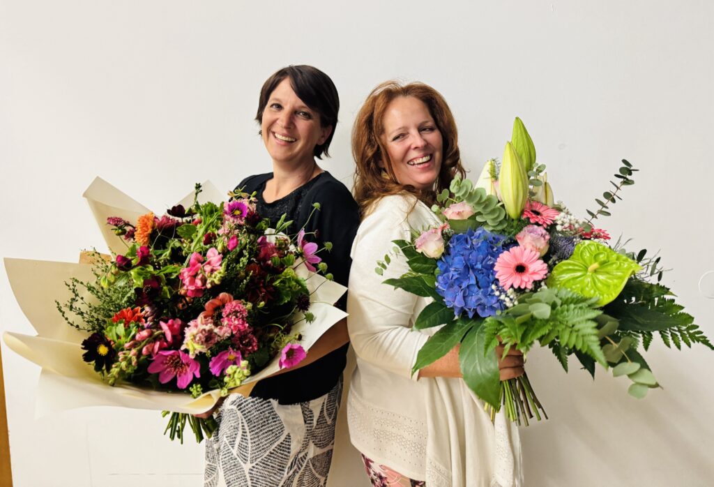 Die Studienkoordinatorinnen Judith Aichbauer-Strauß und Marion Verdorfer stehen nebeneinander und halten einen großen, farbenfrohen Blumenstrauß in den Händen.