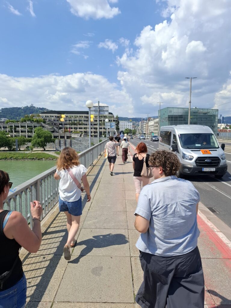 Personen gehen bei sonnigem Wetter über eine Brücke entlang eines Flusses, während rechts Fahrzeuge auf der Straße fahren und moderne Gebäude im Hintergrund zu sehen sind.